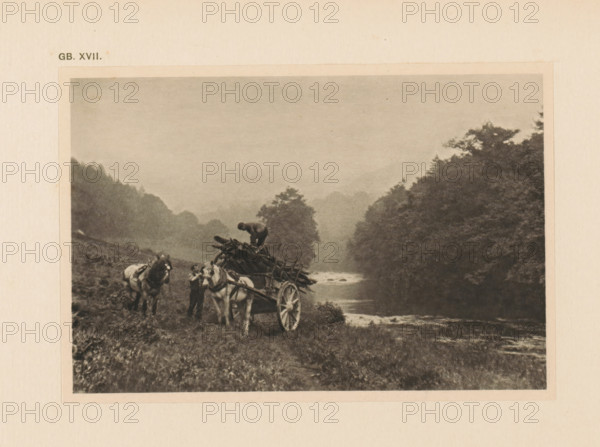 Landscape with horse and carriage, page, photomechanical print, inscription: 'GB. XVII.', inscription: 'Landscape by Edward Hepburn', photographer: Edward Hepburn, cliché maker: anonymous, c. 1900 - in or before 1905, paper, autotypie, height 94 mm x width 141 mm, four-wheeled, animal-drawn vehicle, eg: cab, carriage, coach, landscapes in the temperate zone (landscape with figures, staffage), A sepia-toned photograph showing a rural riverside scene with two horses hitched to a wooden cart loaded with logs, a person standing beside the horses, and another person perched atop the pile of logs; the foreground has grassy terrain, the middle ground shows a winding river with ripples, and the background features tree-covered hills and a misty sky.