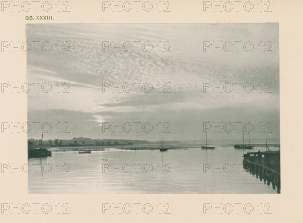 Water landscape with sailing boats, page, photomechanical print, inscription: 'Land & sea by WJ Day', inscription: 'GB. XXXIII.', photographer: William James Day, cliché maker: anonymous, c. 1900 - in or before 1905, paper, autotypie, height 97 mm x width 144 mm, sailing-ship, sailing-boat, landscapes with waters, waterscapes, seascapes (in the temperate zone), A grayscale scene showing a calm body of water with reflections, small boats moored or floating, and vertical masts rising from some of the boats, a distant shoreline with low tree silhouettes, a pier or jetty extending into the water on the right with visible posts and railings, and a sky filled with layered clouds and streaks of light near the horizon.