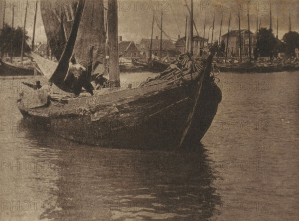 Sailboat setting sail with the sail being hoisted, page, photomechanical print, inscription: 'GB. XXXIV.', inscription: 'Hauling sail by Arthur Marshall', photographer: Arthur Marshall, cliché maker: anonymous, c. 1900 - in or before 1905, paper, autotype, height 114 mm x width 154 mm, sailing-ship, sailing-boat (view, vehicle, ship, etc.), A sepia-toned photograph showing a close view of a wooden sailboat floating on calm water with its bow angled toward the viewer, a coiled sail and ropes piled on the deck near the bow, a partially raised sail and mast amidships, figures on the boat working with the rigging, reflections of the boat and masts in the water, and a distant shoreline lined with more masts and several buildings silhouetted against the sky.