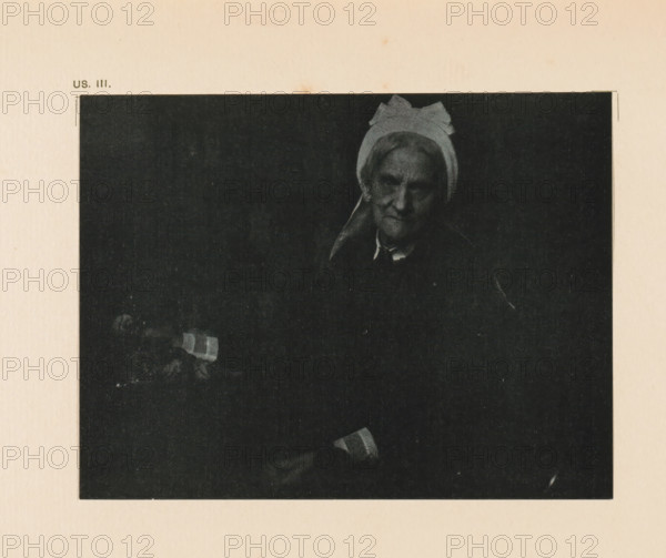 Portrait of an unknown old woman, page, photomechanical print, inscription: 'Portrait of an old lady by Gertrude Käsebier', inscription: 'US. III.', photographer: Gertrude Käsebier, cliché maker: anonymous, c. 1900 - in or before 1905, paper, autotypie, height 110 mm x width 144 mm, historical persons - BB - woman, old woman, A dark, rectangular photograph centered on a pale textured background shows an older woman wearing a light-colored head covering with a bow-like shape at the top and a dark garment, her face partially illuminated against a black backdrop, with one hand visible near the bottom holding a small striped object.