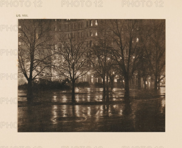 Street view at night, page, photomechanical print, inscription: 'US. VIII.', inscription: 'Reflections night by Alfred Stieglitz', photographer: Alfred Stieglitz, cliché maker: anonymous, c. 1900 - in or before 1905, paper, two-color autotype, height 109 mm x width 142 mm, street, night, A dark, grainy scene shows several leafless trees with bare branches silhouetted against a softly illuminated background, their trunks reflected in a wet, glistening surface below; behind the trees faint, blurred shapes of a multi-story building appear with small bright rectangles of light, and the overall image has a muted, sepia-toned appearance.