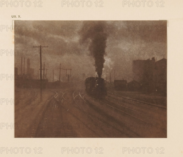 Railway lines with steam train, page, photomechanical print, inscription: 'The hand of man by Alfred Stieglitz', inscription: 'US. X.', photographer: Alfred Stieglitz, cliché maker: anonymous, c. 1900 - in or before 1905, paper, two-color autotype, height 109 mm x width 142 mm, railway, train, A sepia-toned image showing a railway scene with tracks curving into the distance, a dark locomotive near the center emitting a tall plume of smoke that rises into a cloudy sky, silhouettes of telegraph poles and industrial structures along the left side, and faint outlines of buildings on the right, all framed by a pale border.