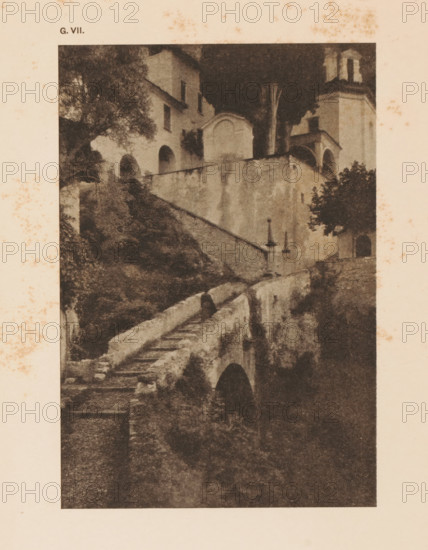 Cityscape in the mountains, page, photomechanical print, inscription: 'Romantic Landscape by Dr. H. Hoek', inscription: 'G. VII.', photographer: H. Hoek, cliché maker: anonymous, c. 1900 - in or before 1905, paper, autotypie, height 148 mm x width 101 mm, city-view in general, 'veduta', A sepia-toned photographic print shows a stone stairway and retaining walls leading up to a cluster of buildings with arched openings and a bell tower. Trees and shrubs grow around the structures, with foliage visible near the building facades and along the stair path. The stone surfaces display weathering and stains, and a narrow arched passage is visible beneath part of the walkway. The image is bordered by a pale margin that has scattered brown spots.