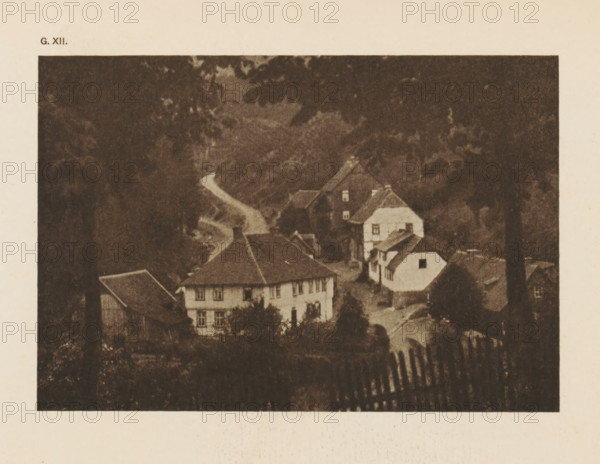 Village view in the Harz Mountains, page, photomechanical print, inscription: 'G. XII.', inscription: 'A Hartz village by HW Müller', photographer: HW Müller, Harz, cliché maker: anonymous, c. 1900 - in or before 1905, paper, autotypie, height 103 mm x width 151 mm, village, mountains, A sepia-toned image of a village scene framed by dark foliage at the top and sides, showing several multi-story houses with sloped roofs and visible windows clustered along a winding road, a small white building with a dark roof set slightly apart, trees and bushes surrounding the buildings, and a foreground area with a fence-like structure appearing as vertical slats.