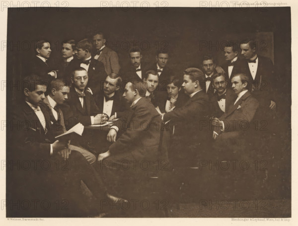 Group of men in suits, including one with a book in hand, page, photomechanical print, photographer: Wilhelm Weimer, Darmstadt, cliché maker: Blechinger & Leykauf, Vienna, c. 1895 - in or before 1900, printer (person), Lower Austria, Hessen, paper, photoaquatint, height 145 mm x width 196 mm, book, adult man (more than eight persons), ensemble of pieces of clothing (men's clothes), A sepia-toned group portrait shows many young men arranged closely together in formal attire, most wearing dark jackets with white shirts and bow ties, seated and standing around a central table; several men hold papers or books, one man looks down at an open book in his hands while another leans forward with hands clasped on the table, faces display neutral expressions, hairstyles are short and neatly combed, a few wear jackets with visible lapels and pocket squares, one man in profile rests an arm on a chair back, another stands with a hand on a seated shoulder, the background is dark and plain, and the im...
