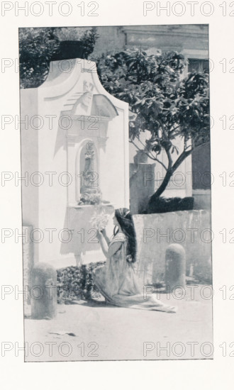 Woman praying in front of a statue of Mary, page, photomechanical print, photographer: Paul Bergon, cliché maker: Jean Malvaux, Brussels, c. 1895 - in or before 1900, paper, autotypie, height 202 mm x width 114 mm, one person praying, the Virgin Mary, A tall rectangular structure with decorative molding and a recessed niche holds a relief plaque and a small ledge with a vase of flowers, positioned next to a leafy tree with a curved trunk; a woman with long dark hair and a flowing dress kneels in front of the structure holding a bouquet of flowers up toward the niche, with a low wall and cylindrical bollards nearby and a paved ground in the foreground.