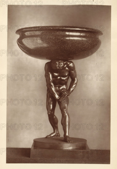 Porphyry bowl supported by a bronze statue of Hercules, from the collection of Christopher Beckett Denison, page, photomechanical print, number: '807', c. 1880 - in or before 1885, paper, Woodburytype, height 186 mm x width 122 mm, Print on seventh unnumbered page after page 78., (story of) Hercules (Heracles), dish, plate, saucer, piece of sculpture (precious metals, arts), A monochrome photograph of a muscular male figure sculpture standing on a round plinth, the figure bent forward with head lowered and hands clasped near the groin, supporting a large shallow bowl atop the shoulders; the sculpture shows glossy highlights and detailed musculature, with one foot placed slightly forward and toes visible, and the bowl has a rounded rim and a speckled surface.