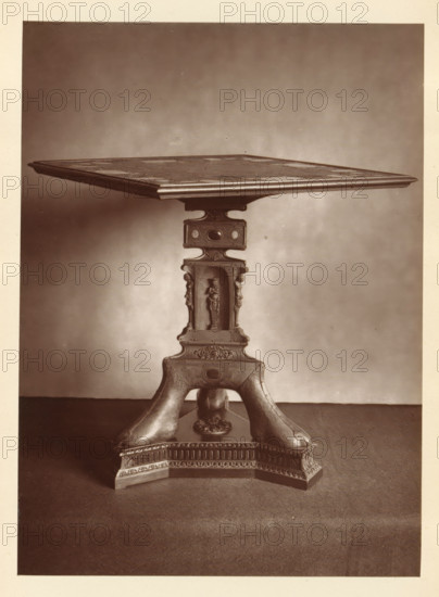 Table in front of a chessboard, from the collection of Christopher Beckett Denison, page, photomechanical print, number: '813', c. 1880 - in or before 1885, paper, Woodburytype, height 171 mm x width 125 mm, Print on fifth unnumbered page after page 80., chess, table (wood and other phytogenic material), A sepia-toned photograph of a decorative wooden table showing the square tabletop with a molded edge and a patterned surface, supported by a carved central pedestal. The pedestal features a rectangular section with rounded cutouts and ornamental oval insets, below which is a recessed niche containing a standing carved figure flanked by vertical decorative elements. The pedestal continues into a sculpted base with curved legs that sweep outward and rest on a molded plinth decorated with carved frieze patterns and a central floral medallion; the surface beneath the table appears to be a textured floor and a plain wall background.