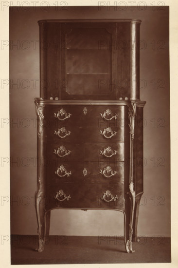 Chest of drawers in the style of Louis XV, from the collection of Christopher Beckett Denison, page, photomechanical print, number: '535', c. 1880 - in or before 1885, paper, Woodburytype, height 195 mm x width 121 mm, ornamental cupboard (wood and other phytogenic material), A tall wooden cabinet with an upper display section featuring a glass-fronted door and interior shelf sits atop a lower chest of drawers. The chest front shows ornate metal drawer pulls and escutcheons arranged symmetrically, with decorative carved appliques at the corners and along the vertical edges. The piece stands on curved, cabriole legs with carved detailing that continues up the sides. The wood surface has a polished finish with visible grain and subtle reflections, and the photograph is in sepia tones against a plain backdrop and floor.