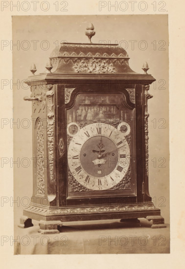 Clock, photograph, page, c. 1874 - in or before 1879, photographer, paper, albumen print, height 144 mm x width 95 mm, A vertically oriented photograph of an ornate wooden clock case standing on a draped surface, showing the upper decorative crest with a finial on top flanked by smaller finials, carved floral and foliate motifs along the cornice and sides, and a central glazed door revealing an inner painted scene above the clock face; the round clock dial has an inner circular plate with ornate hands and an outer ring marked with hour indicators and decorative spandrels, while the surrounding wood surfaces feature carved pilasters, applied scrollwork, and molded base with carved apron and bracket feet.