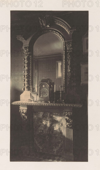 Chimney with clock in the house of Antonin Terme, photograph, page, Liège, c. 1880 - in or before 1885, photographer, paper, platinum process, height 212 mm x width 112 mm, clock, art collection, art gallery, chimney-piece, mantel piece, A dark photograph of an ornate mantel and mirror with heavy carved decoration framing an arched mirror, a decorative clock sitting on the mantel flanked by two candlesticks, patterned wallpaper and framed pictures visible in the reflected and surrounding walls, and sculpted floral and acanthus motifs on the mantel and pilasters beneath the shelf.