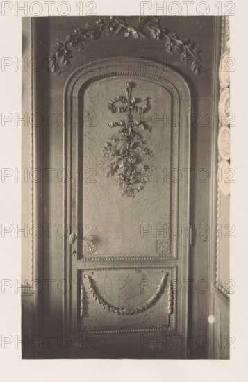 Wooden decorated door in the house of Antonin Terme, photograph, page, Liège, c. 1880 - in or before 1885, photographer, paper, platinum process, height 215 mm x width 132 mm, door, art collection, art gallery, sculpture (relief, sculpture), A tall wooden panel with an arched top is shown, featuring carved decorative motifs including a bow with hanging foliage near the upper center, ornate garlands draped across a lower recessed panel, and carved floral rosettes at the corners of a rectangular frame; the panel displays textured wood grain and raised moulding along its borders, with portions of adjacent wall and a series of decorative plates visible along the right edge.