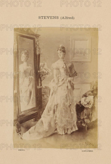 Stevens (Alfred), Photo reproduction of Jeune femme à sa toilette by Alfred Stevens, photograph, reproduction, page, photographer: Alexandre, after painting by Alfred Stevens, c. 1888 - in or before 1893, paper, albumen print, albumendruk, height 149 mm x width 105 mm, adolescent, young woman, maiden, looking in the mirror, contemplation of own beauty, painting, drawing and the graphic arts, A sepia-toned portrait of a woman in an elaborate gown standing beside a tall ornate mirror, the mirror reflecting her profile; she holds a folded fan at her side and wears a ruffled bodice with off-the-shoulder sleeves, and her hair is styled up with decorative accents; a framed picture hangs on the wall behind her and a cushioned chair with a patterned fabric sits to her side, while a small floral arrangement is visible near the mirror.