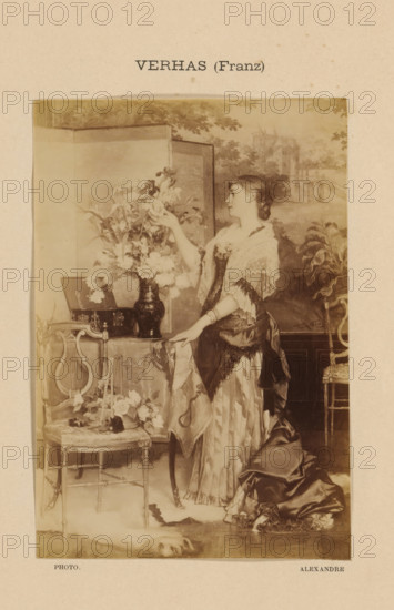 Photo reproduction of Le bouquet by Frans Verhas, Verhaz (Franz), photograph, reproduction, page, photographer: Alexandre, after painting by Frans Verhas, c. 1888 - in or before 1893, paper, albumen print, albumendruk, height 147 mm x width 101 mm, flowers, adolescent, young woman, maiden, painting, drawing and the graphic arts, A vertically oriented sepia photographic print shows a young woman standing beside a small table draped with a patterned cloth, reaching toward a large vase of flowers placed on the table; she wears a layered dress with a shiny skirt and a lace shawl over her shoulders, her hair pulled back in a braided style, and one hand rests on the table while the other holds a flower. The scene includes ornate chairs with carved details, a small framed item leaning on the table, scattered blossoms on the chair seat and floor, and a decorative folding screen or painted backdrop behind the woman depicting trees and architectural forms. The image is mounted on a textured ligh...