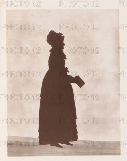 Photo reproduction of a drawing depicting a silhouette of Anna Maria Shipley, reproduction, page, photomechanical print, cliché maker: Woodbury Permanent Photographic Printing Company, after drawing by anonymous, c. 1866 - in or before 1876, photographer, paper, Woodburytype, Woodburytypie, height 118 mm x width 91 mm, silhouette-apparatus, 'physionotrace', A side silhouette of a standing person wearing a long dress and a hat with decorative elements, holding a small rectangular object in one gloved hand, with visible shoes at the hem, all set against a plain, lightly textured background.