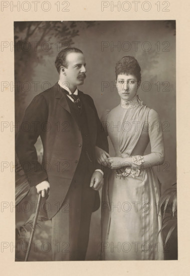 The Duke and Duchess of Fife, Portrait of Alexander Duff and his wife Louise Victoria Alexandra Dagmar of Saxe-Coburg and Gotha, page, photomechanical print, inscription: '57 & 61, Elbury Street, London.', photographer: Downey, William & Daniel, London, clichémaker: anonymous, c. 1880 - in or before 1890, paper, Woodburytype, Woodburytypie, height 139 mm x width 94 mm, Person, Louise Victoria Alexandra Dagmar (Princess of the United Kingdom), Alexander William George Duff (Duke of Fife), historical persons, historical persons - BB - woman, A sepia-toned portrait shows a standing man and woman positioned close together against a plain backdrop with faint foliage shadows; the man wears a dark three-piece suit with a high-collared shirt and tie, has short hair and a mustache, and holds a cane in one hand while his other hand rests near his waist; the woman wears a light-colored, high-necked dress with pleated and ruffled details at the bodice and cuffs, has hair styled up, and clasps her ...