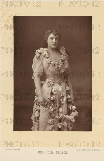 Portrait of Julia Neilson in a dress with flowers, Miss Julia Neilson, page, photomechanical print, inscription: '57 & 61, Elbury Street, London.', photographer: Downey, William & Daniel, London, cliché maker: anonymous, c. 1882 - in or before 1892, paper, Woodburytype, height 140 mm x width 95 mm, Person, Julia Neilson, portrait of actor, actress, A sepia studio portrait of a young woman standing and facing forward, wearing a light, flowing gown decorated with clusters of flowers around the neckline, sleeves, waist, and cascading down the skirt; her hair is wavy and pulled back from her face, she has a neutral facial expression and is looking slightly to the side, her bare arms are visible and her hands rest at her sides holding part of the floral garlands, the background is plain and dark, and the photograph is mounted on a light-colored card with some small spots and foxing visible around the edges.