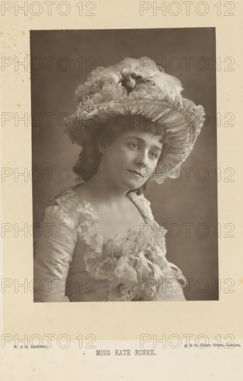 Miss Kate Rorke, Portrait of Kate Rorke, page, photomechanical print, inscription: '57 & 61, Elbury Street, London.', photographer: Downey, William & Daniel, London, cliché maker: anonymous, c. 1882 - in or before 1892, paper, Woodburytype, height 140 mm x width 95 mm, Person, Kate Rorke, portrait of actor, actress, A sepia portrait of a woman shown from the chest up wearing a wide, ruffled hat decorated with layered fabric and flowers, with curly hair visible beneath the brim; she has a soft, neutral facial expression and is turned slightly to one side; her dress has lace trim around the neckline and bodice and features bows and gathered fabric; the background is plain and evenly shaded and the portrait is mounted on a bordered card with visible aging spots.
