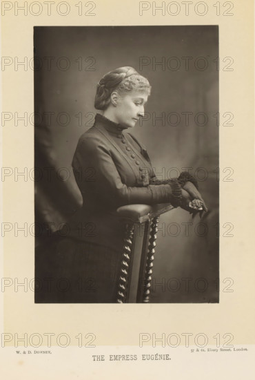 The empress Eugénie, Portrait of Eugénie de Montijo, page, photomechanical print, inscription: '57 & 61, Elbury Street, London.', photographer: Downey, William & Daniel, London, cliché maker: anonymous, c. 1882 - in or before 1892, paper, Woodburytype, height 140 mm x width 93 mm, Person, Eugénie (Empress of the French), ruler, sovereign - BB - female ruler, A sepia-toned portrait of a seated woman shown in profile looking downward, wearing a dark high-collared dress with a row of fabric-covered buttons down the front and decorative ruffled cuffs, her hair covered by a textured cap, hands resting on the padded arm of a turned-wood chair while holding a small cluster of flowers, a softly blurred backdrop behind her and a wide pale margin framing the photograph.