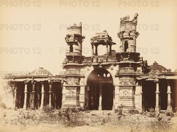 Mulik Alum's Mosque, View of the Malik Alam Mosque in Ahmedabad, photograph, page, number: '10.', inscription: 'D2', photographer: Thomas Biggs, Ahmedabad (city), c. 1856 - in or before 1866, Thomas Biggs, photographer, Gujarat, paper, albumen print, height 149 mm x width 201 mm, temple, shrine, Islam, Mohammedanism, Malik Alam Mosque, An ornate stone gateway with two cylindrical towers flanking a central arched entrance is shown, featuring carved decorative bands and sculptural details on the towers and surrounding walls; a small pavilion-like structure sits above the arch supported by columns, and domed roofs and colonnaded walkways extend to either side; the foreground shows sparse vegetation and rough ground leading up to the base of the structure, and the image has a high contrast, sepia-toned appearance.