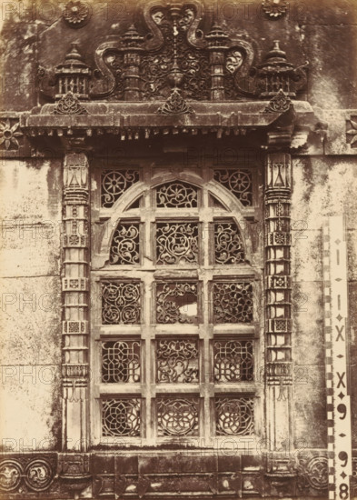 Window of the Shapoor Mosque in Ahmedabad, The Shápoor Mosque.- Window of perforated stone, photograph, page, number: '111.', photographer: Thomas Biggs, Ahmedabad (city), c. 1856 - in or before 1866, Gujarat, paper, albumen print, height 188 mm x width 136 mm, temple, shrine, Islam, Mohammedanism, window, A sepia photograph shows an ornate stone window set into a textured wall, framed by carved vertical columns with geometric and floral motifs and a decorative lintel above featuring scrollwork and small pavilion-like structures; the window itself has an arched top and is divided into a grid of panels filled with intricate pierced stone latticework in swirling and floral patterns, with areas of weathering and darkened patches on the surrounding stone, and a patterned measuring strip placed vertically beside the right side of the window.