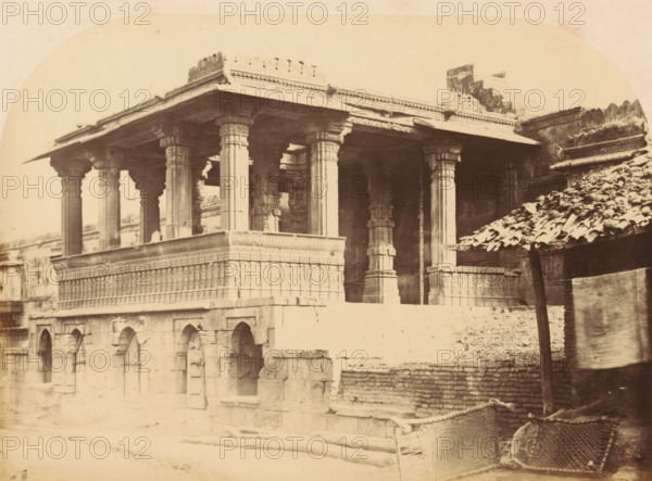 The Jumma Mosque.- Hindoo Porch in the center of the Southern Colonnade, Portal in a colonnade of the Jama Masjid in Ahmedabad, photograph, page, number: '14.', inscription: 'E2', photographer: Thomas Biggs, Ahmedabad (city), c. 1856 - in or before 1866, Gujarat, paper, albumen print, height 135 mm x width 188 mm, colonnade, temple, shrine, Islam, Mohammedanism, monumental door, porch, 'aediculum', Jama Masjid (Ahmedabad), A sepia-toned photograph shows a stone structure with an elevated open porch supported by carved columns and ornate capitals, a richly decorated frieze running along the top edge, and a patterned balustrade beneath the porch; beneath and beside the porch are arched recesses set into a lower stone wall, and to the right a sloped roof covered with tiles leans on a wooden post above a pile of stones and woven baskets.