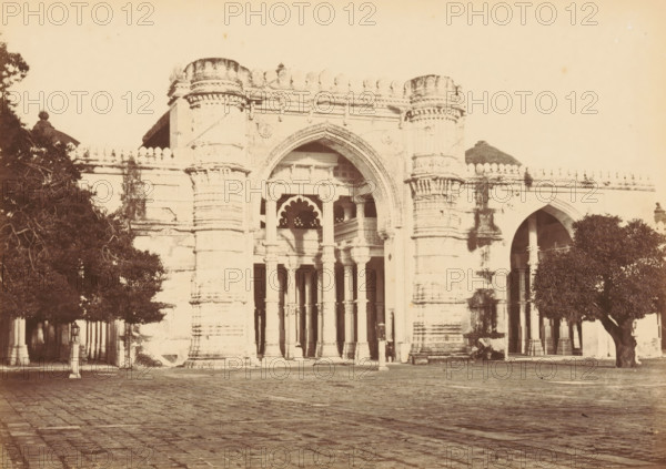 Façade of the Jama Masjid in Ahmedabad, The Jumma Mosque.- Nearer view, photograph, page, number: '15.', photographer: Thomas Biggs, Ahmedabad (city), c. 1856 - in or before 1866, Gujarat, paper, albumen print, height 136 mm x width 189 mm, temple, shrine, Islam, Mohammedanism, parts of church exterior and annexes: facade, Jama Masjid (Ahmedabad), A sepia-toned photograph of a large ornate stone gateway with a central pointed arch flanked by tall cylindrical columns with decorative horizontal bands, a series of slender columns and scalloped arches visible within the entrance, and carved architectural detailing along the top edge; trees frame both sides of the gateway, casting shadows on the facade and standing beside a paved courtyard in the foreground.