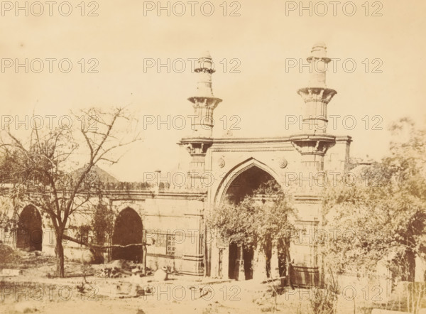 Ahmed Shah's Mosque, View of Ahmed Shah's Mosque in Ahmedabad, photograph, page, number: '2.', photographer: Thomas Biggs, Ahmedabad (city), c. 1856 - in or before 1866, Gujarat, paper, albumen print, height 136 mm x width 184 mm, temple, shrine, Islam, Mohammedanism, Ahmed Shah Mosque, A sepia-toned photograph showing a stone gateway with an arched central entrance flanked by decorative columns and two tall cylindrical towers rising above the structure, leafless trees and foliage partially obscuring the lower sections of the building, a series of smaller arches along the facade to the left, and scattered stones and ground-level debris in the foreground.