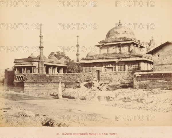 View of Rani Sipri's Mosque and a tomb in Ahmedabad, Ránee Seeprees Mosque and Tomb, photograph, page, number: '21.', inscription: 'G', photographer: Thomas Biggs, Ahmedabad (city), c. 1856 - in or before 1866, Thomas Biggs, photographer, Gujarat, paper, albumen print, height 144 mm x width 195 mm, temple, shrine, Islam, Mohammedanism, grave, tomb, Rani Sipri Mosque, A wide sepia photograph of an ornate stone complex featuring a large domed structure with a smaller dome cluster to its left, decorative cornices and carved window screens, flanked by tall slender minaret-like towers with stacked cylindrical segments, and a colonnaded veranda supported by carved pillars; a low brick wall runs along the front with a narrow doorway set into it and rubble and uneven ground in front, while a simple building with a sloped roof appears at the right edge and a mostly clear sky fills the background.