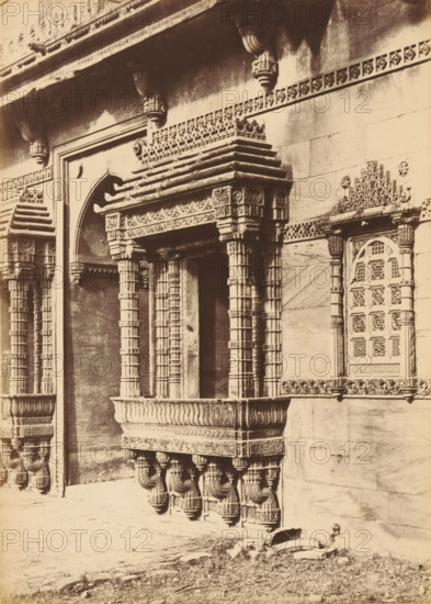 Façade of the Rani Rupamati Mosque in Ahmedabad, The Queen's Mosque in Mirzapoor.- The Northern Side-arch and Windows, photograph, page, number: '31.', photographer: Thomas Biggs, Ahmedabad (city), c. 1856 - in or before 1866, Thomas Biggs, photographer, Gujarat, paper, albumen print, height 188 mm x width 135 mm, temple, shrine, Islam, Mohammedanism, parts of church exterior and annexes: facade, Rani Rupamati Mosque, A sepia photograph of a stone building façade featuring ornate carved columns, brackets, and decorative ledges forming a balcony-like projection, with intricately patterned lattice window panels set into the wall; the carved elements include layered cornices, geometric and floral motifs, and sculpted supports beneath the projecting platform; a framed arched recess appears beside the central projection, and a patterned frieze runs horizontally across the upper wall; the ground in front shows rough stone and sparse grass.