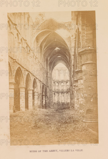 Ruins of the abbey, Villers la ville, photograph, page, Villers-la-Ville, c. 1856 - in or before 1866, photographer, paper, albumen print, height 177 mm x width 118 mm, ruin of church, monastery, Villers Abbey, The image shows a faded, sepia-toned interior view of a large ruined stone building with tall vaulted arches and columns lining both sides, a long central nave leading toward a distant apse with multiple arched openings, broken roof sections revealing sky, rough stone surfaces with visible weathering and chipped areas, overgrown vegetation covering the floor, and scattered rubble along the ground.