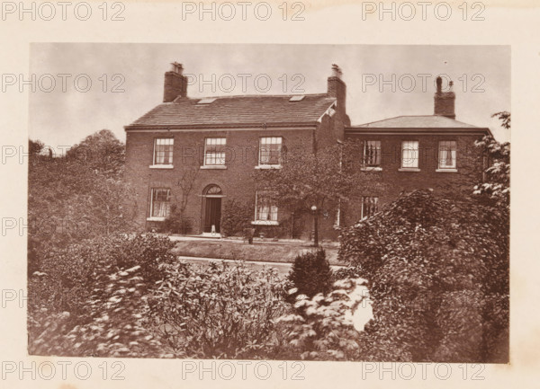Exterior of John Bright's birthplace at Rochdale, page, photomechanical print, Rochdale, c. 1873 - in or before 1878, photographer, printmaker, Manchester, paper, Woodburytype, height 77 mm x width 118 mm, housing, façade (of house or building), A sepia-toned photograph of a two-story brick house with a tiled roof and multiple chimneys, framed by dense shrubs and trees in the foreground and sides. The house shows several sash windows with visible panes on both floors, an arched central doorway with a closed door, and climbing plants against the facade. A path or driveway runs in front of the house, and trimmed hedges and garden plants occupy the foreground. The sky above the house appears cloudy.