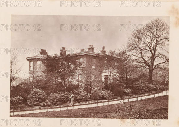 Exterior of (presumably) John Bright's house at Rochdale, page, photomechanical print, Rochdale, c. 1873 - in or before 1878, photographer, printmaker, Manchester, paper, Woodburytype, height 77 mm x width 118 mm, housing, façade (of house or building), A sepia-toned photograph shows a large multi-story brick house partially obscured by dense shrubbery and leafless trees, with a rounded bay window visible at one corner and several chimney stacks rising from the roof; a paved path with a low metal railing runs across the foreground along a grassy slope, and a person wearing light clothing is walking on the path while holding a long tool; a tall bare tree stands to the right of the house and the sky above is pale and empty.