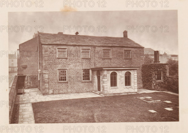 Exterior of a house at Rochdale, page, photomechanical print, Rochdale, c. 1873 - in or before 1878, photographer, printmaker, Manchester, paper, Woodburytype, height 77 mm x width 118 mm, housing, façade (of house or building), A stone building with a pitched slate roof fills the frame, showing a rectangular facade of evenly coursed masonry with several sash windows and a downpipe running vertically; a small single-story projection with arched windows and a doorway extends from the front, and a paved path and low stone wall border a lawn area in the foreground, while an adjacent wall covered in climbing plants and a chimney are visible to one side and rooftops of other buildings appear in the background.