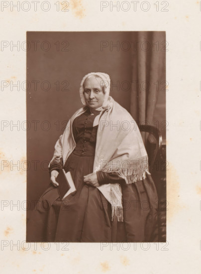 Studio portrait of Hannah S. Allen, page, photomechanical print, c. 1874 - in or before 1884, photographer, printmaker, paper, Woodburytype, height 90 mm x width 59 mm, Print on frontispiece., Person, Hannah S. Allen, historical persons - BB - woman, A seated older woman wearing a dark dress and a light shawl with fringe is shown from head to mid skirt, her hair covered by a lace-trimmed bonnet, holding a closed book in one hand while the other rests on her lap, a dark chair back is visible behind her and a plain curtain hangs to one side, the photograph is bordered by a wide light-colored mount with scattered small stains.