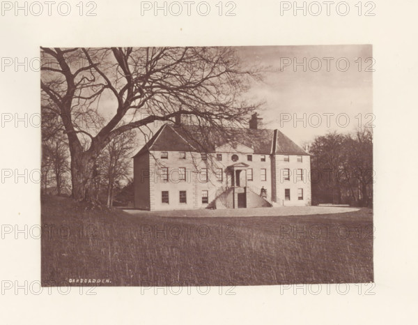 Garscadden, photograph, page, photographer: Thomas Annan, Glasgow, c. 1868 - in or before 1878, paper, carbon print, height 115 mm x width 160 mm, rural housing, eg country-house, villa, cottage, façade (or house or building), A large pale building with a central staircase and a portico is shown set on a grassy foreground, bare-branched trees frame the scene with a very large tree on the left whose branches extend across the sky, the building has multiple windows and a pitched roof with a chimney, a person stands on the staircase near the entrance, and trees and shrubbery are visible behind and to the sides of the building.