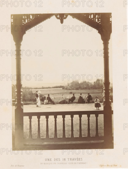 Company in front of the kiosk on the Plateau de Gravelle, Une des 16 travées du kiosque de Gravelle (côté de Pontenay), Bois de Vincennes, photograph, page, photographer: Ildefonse Rousset, Plateau de Gravelle, c. 1861 - in or before 1866, Paris, paper, albumen print, height 163 mm x width 123 mm, wood and park (as places of recreation), summer-house, arbour, garden-house (Dutch: 'tea dome'), A sepia photograph framed by ornate wooden posts and a carved roof edge shows a group of people seated at outdoor tables on a lawn, with a low fence and distant trees in the background; several seated figures wear long coats or dresses and hats, one standing figure in a uniform or apron walks near the group, and a small child stands closer to the foreground; a wooden balustrade fills the lower foreground, and the scene is lit with soft, even daylight.