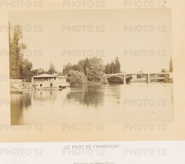 View of the Pont de Champigny in Champigny-sur-Marne, Le pont de Champigny, photograph, page, photographer: Ildefonse Rousset, Île-de-France, c. 1860 - in or before 1865, paper, albumen print, height 123 mm x width 178 mm, river, bridge, Marne (river), Pont de Champigny, Champigny-sur-Marne, A sepia-toned riverscape showing a calm stretch of water reflecting the sky and surrounding features, with a low two-story riverside building with a covered balcony and small boats moored beside it at the near left, dense clusters of trees rising behind and along the riverbank toward the center, and an arched bridge with decorative supports crossing the river toward the right, with more trees and grassy banks visible beyond the bridge.