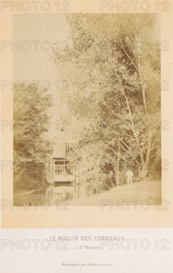 Le moulin des Corbeaux à St. Maurice, Exterior of the Moulin des Corbeaux in Saint-Maurice, photograph, page, photographer: Ildefonse Rousset, Saint-Maurice (Val-de-Marne), c. 1860 - in or before 1865, paper, albumen print, height 175 mm x width 133 mm, river, watermill in landscape, façade (of house or building), Marne (river), A faded photographic print showing a tall stand of leafy trees on the right and a water channel running toward the foreground, with dense foliage on the left and a partially visible wooden building or mill structure over the water near the center; a person stands on the grassy bank at right holding a parasol and facing the water; reflections of the structure and trees appear in the water; the image is mounted on a larger cream mount with printed text below the photograph.