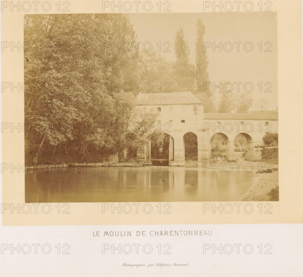 Le moulin de Charentonneau, Exterior of the Moulin Brûlé in the Marne, photograph, page, photographer: Ildefonse Rousset, Île-de-France, c. 1860 - in or before 1865, paper, albumen print, height 128 mm x width 173 mm, river, watermill, façade (of house or building), Marne (river), A sepia-toned photograph showing a riverside scene with a stone building spanning the water on large arched supports, trees and foliage clustered to the left and behind the building, reflections of the arches and shoreline visible in the calm river, a small wooden dock and a few small boats along the near bank, and a narrow strip of sandy shore on the right.