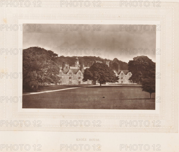 Knole House, View of Knole House, page, photomechanical print, photographer: Stephen Thompson, Kent, c. 1866 - in or before 1876, printmaker, paper, Woodburytype, height 102 mm x width 160 mm, country-house, A sepia-toned photograph framed by an ornate embossed border shows a large stone building with multiple chimneys and gabled rooflines partially obscured by mature trees; a wide expanse of lawn stretches in front of the building with a single small figure seated near the center, and a gravel path runs parallel to the facade beneath a cloudy sky.