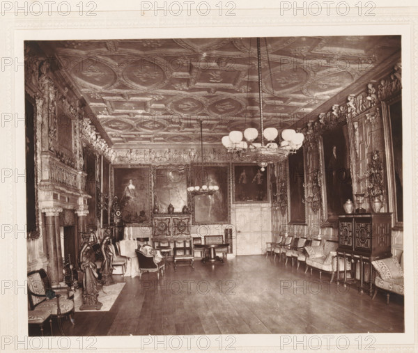 The ball room, Knole, Interior of the ballroom of Knole House, page, photomechanical print, photographer: Stephen Thompson, Knole House, c. 1866 - in or before 1876, printmaker, Kent, paper, Woodburytype, Woodburytypepie, height 148 mm x width 187 mm, interior of the house, A large ornate room with a high decorated ceiling featuring patterned panels and ornamental motifs, two hanging chandeliers with round lamps suspended by long chains, carved and sculpted wall moldings along the upper walls, several large framed paintings mounted on the walls, an elaborate fireplace with columns and carved figures on one side, rows of upholstered chairs and small tables arranged along the walls, decorative cabinets and chests with ornate inlay and vases on top, sculpted statues and carved wooden figures placed near the fireplace and along the walls, patterned cushions on the seating, a polished wooden floor, and various decorative metal sconces and ornamental flourishes throughout the space.