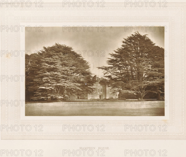View of Hampden House, Hampden House, page, photomechanical print, photographer: Stephen Thompson, Great and Little Hampden, c. 1866 - in or before 1876, printmaker, Buckinghamshire, paper, Woodburytype, height 102 mm x width 160 mm, country-house, A sepia-toned photograph mounted on a decorative mat shows a large stately building partially obscured by tall, dense trees with spreading branches on either side, a broad lawn in the foreground, and a cloudy sky above.