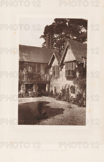 Gables, Ightham Mote, View of the Ightham Mote near Ightham, page, photomechanical print, photographer: Stephen Thompson, Ightham, c. 1866 - in or before 1876, printmaker, Kent, paper, Woodburytype, height 160 mm x width 102 mm, courtyard, rural housing, eg country-house, villa, cottage, A sepia-toned photograph shows a courtyard framed by a timber-framed building with steep gabled roofs and leaded lattice windows, climbing plants on the walls, and a stone lower façade with an arched window; a tall chimney rises behind the roofline and large trees fill the background; the cobbled courtyard contains a decorative urn on a plinth casting a shadow and a small metal chair near the building, with a shaded recess under the eaves where foliage and a small table are partially visible.
