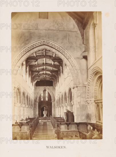 Walsoken, photograph, page, photographer: Edward Johnson, All Saints' Church (Walsoken), c. 1858 - in or before 1869, Edward Johnson, photographer, Cambridgeshire, paper, albumen print, height 178 mm x width 126 mm, interior of church, facade (of house or building), A sepia photograph showing the interior of a church nave with a central aisle flanked by wooden pews and carved choir stalls, a patterned stone floor leading toward a raised pulpit or font in the distance, tall arched openings and columned arcades along the side walls, ornate stone archways with decorative moldings, a timber roof with exposed beams and trusses, high walls with plastered surfaces, stained glass windows visible at the far end, and various stone and wooden architectural details and furnishings along the sides.