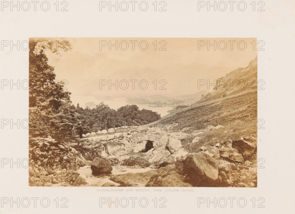 Derwentwater and Skiddaw, from Ashness Bridge, View of Derwentwater and Mount Skiddaw in the Lake District, photograph, page, photographer: Garnett & Sproat (possibly), Lake District, c. 1857 - in or before 1871, paper, albumen print, height 107 mm x width 164 mm, lake, mountains, Derwentwater, Skiddaw, A sepia-toned landscape photograph showing a rocky hillside and stream in the foreground with a stone arch bridge spanning the water and scattered boulders and fallen logs nearby, a person wearing a hat standing beside a wooden cart or gate near a rocky path, dense trees and shrubs on the left, a sloping grassy hillside rising on the right, and a distant view of misty hills and a large body of water beyond the tree line under a pale sky.