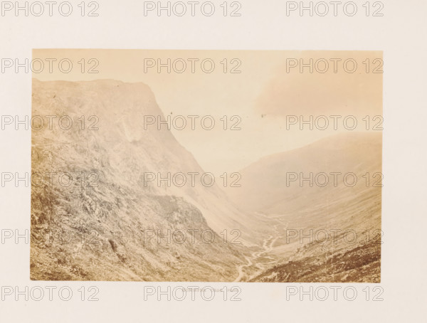 Honister Crag Pass, View of Honister Pass in the Lake District, photograph, page, photographer: Garnett & Bowers, Lake District, c. 1857 - in or before 1870, paper, albumen print, height 109 mm x width 164 mm, dale, valley, Honister Pass, A sepia-toned photograph of a steep rocky valley with a rugged cliff face on the left and sloping hills on the right, a winding narrow stream running through the valley floor, textured rock surfaces and scree visible on the slopes, a pale sky above with faint cloud shapes, and a wide light border surrounding the image.