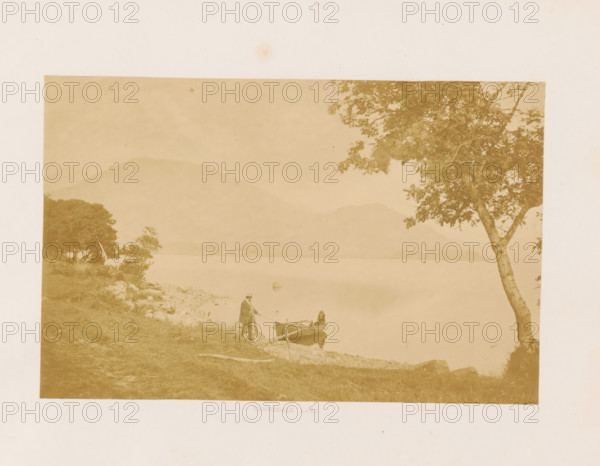 View of Ennerdale Water in the Lake District with a rowboat at the shore, Ennerdale Lake, photograph, page, photographer: Garnett & Bowers, Lake District, c. 1857 - in or before 1870, paper, albumen print, height 106 mm x width 164 mm, lake, mountains, Cumbria, Ennerdale Water, A sepia-toned photograph showing a calm lake with distant hills and a shoreline in the foreground, a small rowboat at the water's edge with a person standing beside it and another seated inside, grassy and rocky ground leading to the shore, clusters of trees on the left and a prominent tree trunk with branches and leaves on the right, and a pale border surrounding the central image.