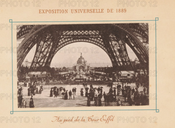 La Tour Eiffel, View of the Eiffel Tower in Paris, Exposition Universelle de 1889, page, photomechanical print, maker: Neurdein Frères, Paris, 1889, paper, collotype, photoprint, height 108 mm x width 69 mm, tower, clock tower, monumental adornment, world fair, world exhibition, World Exhibition 1889 (Paris), Eiffel tower, A sepia-toned photograph framed by a thin blue border shows the underside of a large iron lattice structure forming a sweeping arch above a crowded open space, with ornate metalwork visible along the arch and intricate cross-bracing in the supporting legs; beneath the arch a domed pavilion with sculptural details and fountains sits at the center of a landscaped area, flanked by trees and additional architectural elements; in the foreground a crowd of people dressed in period clothing, many holding umbrellas and wearing long coats and hats, are walking and standing on a paved area, with smaller figures and grouped clusters extending toward the pavilion and the edges o...