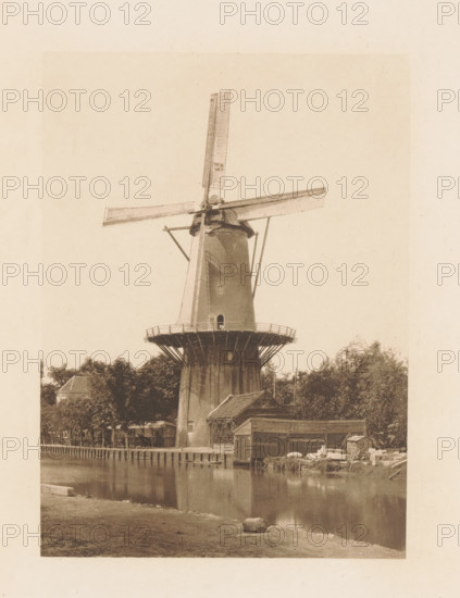 Windmill in the Netherlands, page, photomechanical print, title: 'A Dutch Windmill.', photographer: Charles L. Mitchell, Netherlands, cliché maker: AW Elson & Company, Boston (Massachusetts), c. 1884 - in or before 1894, North America, Massachusetts, paper, photoaquatint, height 121 mm x width 87 mm, Print on frontispiece., windmill in landscape, A tall traditional windmill with large cross-shaped sails rises above a circular wooden gallery and a tapered brick base, surrounded by trees and nearby buildings, with a calm canal reflecting the structures and a grassy bank in the foreground.
